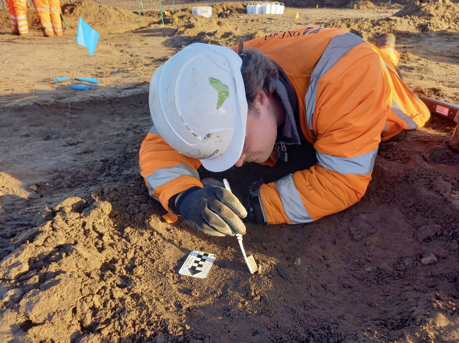 Hadley Sharman, in a hard hat and hi-vis jacket, lying on the ground of an excavation and cleaning a coin with a brush