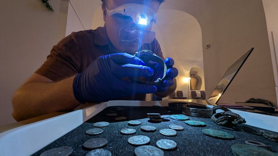 Hadley Sharman, wearing a head loupe and gloves, examining a piece of jewellery above a tray of coins from the Fenwick treasure